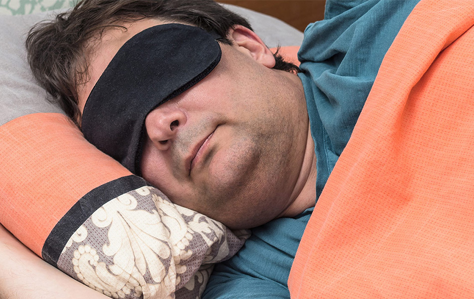 Man sleeping with an orange blanket and wearing a sleep mask.